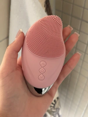 Rosa ansiktsrengöringsborste silikon - Elektrisk ansiktsrengöringsborste i mjuk rosa silikon med små borst för djuprengöring. Har tre knappar på framsidan för olika inställningar och en ergonomisk form som ligger skönt i handen. Perfekt för daglig hudvårdsrutin.