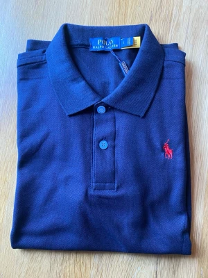 Mörkblå piké från Polo Ralph Lauren - Klassisk mörkblå pikétröja från Polo Ralph Lauren med röd broderad logga på bröstet. Tröjan har krage, knappar framtill och är tillverkad i mjuk bomull. Perfekt för en clean och stilren look.