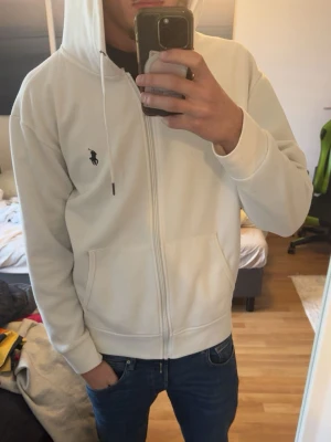 Vit hoodie från Polo Ralph Lauren - Säljer en stilren vit hoodie från Polo Ralph Lauren med dragkedja framtill och klassisk logga på bröstet. Hoodien har huva med dragsko och två fickor framtill. Perfekt för en clean och avslappnad look.