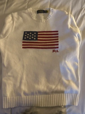Vit stickad tröja Polo Ralph Lauren - Vit stickad tröja från Polo Ralph Lauren med amerikansk flagga i blått, rött och vitt på bröstet samt RL-broderi. Tröjan har rund halsringning och ribbade muddar vid ärmslut och nederkant. Klassisk och stilren design. M passar mer som S typ