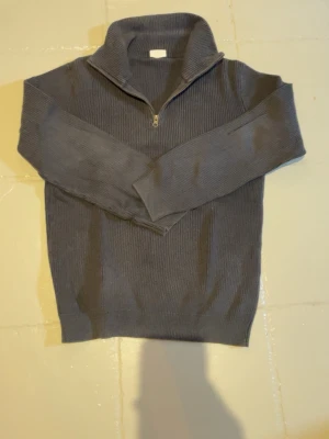 John Henric stickad halvzip tröja - Mysig blå stickad tröja med halvzip och hög krage. Tröjan har ribbad struktur och långa ärmar, perfekt för kyliga sommarkvällar. Enkel och stilren design som passar till det mesta.