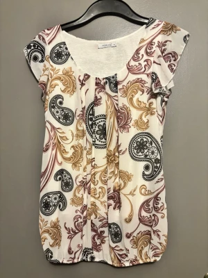 Mönstrad blus från Hailys (köpt utomlands) - Snygg blus från Hailys med mönster i svart, guld och vinrött på vit botten. Blusen har rund halsringning, korta ärmar och lätt rynkad nederkant. Tillverkad i ett mjukt och luftigt material som passar perfekt till jeans eller kjol.