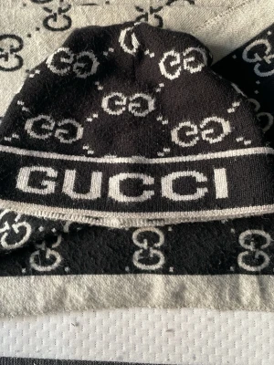 Svartvit halsduk från Gucci plus mössa - Säljer en svartvit halsduk från Gucci med det klassiska GG-mönstret över hela tyget. Halsduken har en kontrasterande design med svart och vitt samt tydlig logga. Perfekt accessoar för dig som gillar lyxiga detaljer och vill sticka ut.