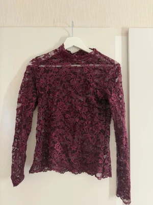 Vinröd spetsblus med lång ärm - Säljer en vinröd, transparent spetsblus med lång ärm och hög hals. Blusen har ett vackert blommigt spetsmönster och är figurnära i modellen. Perfekt för dig som vill ha en trendig och elegant look.