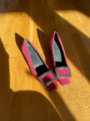 Roger Viver Vinröda mockapumps med fyrkantig detalj - Säljer ett par vinröda pumps i mocka med fyrkantig tå och en stor dekorativ metallspänne framtill. Skorna har låg klack och en elegant, retroinspirerad look. Perfekta för dig som vill sticka ut med stilrena detaljer.
