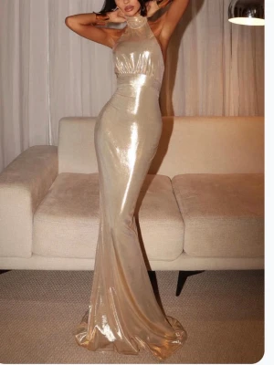 Guldig balklänning med halterneck - Snygg guldig långklänning i glansigt material med halterneck och figurnära passform. Aldrig använd från VioletAurora! Klänningen har öppen rygg och markerad midja, vilket ger en elegant siluett. Perfekt för dig som vill sticka ut på bal eller fest.