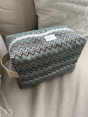 Zigzag-mönstrad necessär från Emelo - Necessär från Emelo i tyg med zigzag-mönster i grönt, blått, beige och svart. Rektangulär form med vitt handtag på sidan och vit dragkedja. Insidan är ljus och rymlig, perfekt för smink eller småprylar. Beige insida med innerfack. Mått ca 27x18x12 cm 