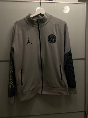PSG x Jordan grå träningsjacka - Snygg grå träningsjacka från PSG x Jordan med svarta detaljer, dragkedja framtill och två fickor. Ärmarna har mörkblått rutmönster och gul text på ena ärmen. Perfekt för sport eller streetwear.