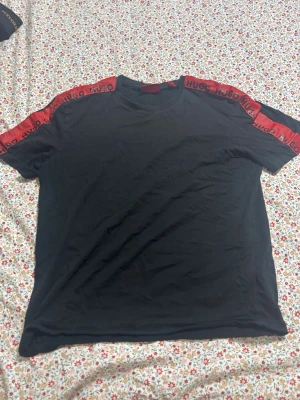 Svart HUGO t-shirt med röda detaljer - Snygg svart t-shirt från HUGO med röda band längs axlarna där det står 'HUGO' upprepat. Klassisk rund halsringning och korta ärmar. Tillverkad i mjuk bomull som känns skön mot huden. Perfekt för en clean och modern look.