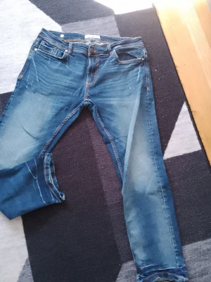 Blå slim fit jeans med slitningar - Säljer ett par blå slim fit jeans med snygga slitningar och tvättade detaljer. Klassisk femficksmodell med orangea sömmar och knappgylf. Perfekta till vardagslooken och passar till det mesta. Materialet är mjukt denim för skön känsla.