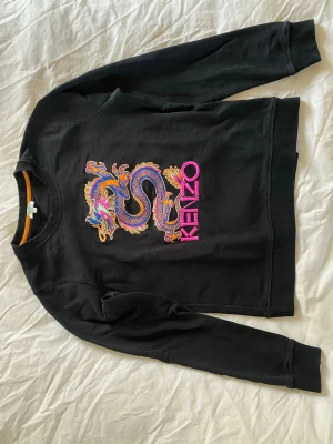 Svart Kenzo sweatshirt med drakmotiv - Unik svart sweatshirt från Kenzo med en stor broderad drake på bröstet och Kenzo-loggan i rosa. Tröjan har rund halsringning och ribbade muddar vid ärmslut och nederkant. Perfekt statement-plagg för dig som gillar unika detaljer. Endast använd ett fåtal gånger