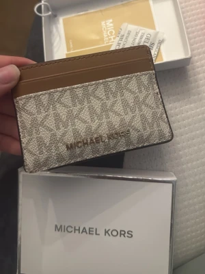 Michael Kors beige kortfodral - Stilrent kortfodral från Michael Kors i beige och brunt med MK-logga över hela framsidan. Tillverkat i canvas med detaljer i brunt läder och guldfärgad logotyp. Perfekt för kort och ID, smidig att ha i fickan eller väskan.