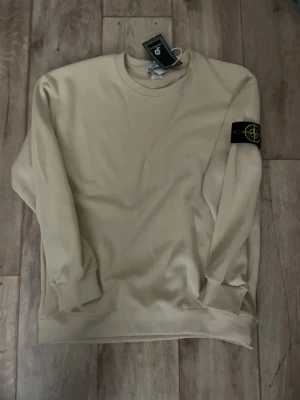 Beige sweatshirt från Stone Island - Snygg beige sweatshirt från Stone Island med klassisk rund hals och ribbade muddar. Tröjan har den ikoniska Stone Island-loggan på vänster ärm och är tillverkad i mjukt bomullsmaterial. Perfekt för en clean och stilren look.