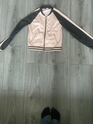 Rosa och svart bomberjacka - Snygg bomberjacka i satinliknande material med ljusrosa kropp och svarta ärmar. Jackan har dragkedja framtill, två fickor med dragkedja och randiga muddar i svart och rosa vid ärmslut och midja. Perfekt för en trendig streetstyle-look.