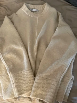 Beige stickad tröja från Zara - Mysig beige stickad tröja från Zara med rund halsringning och breda ribbade muddar vid ärmslut och nederkant. Tröjan har en rak passform och dekorativ söm framtill och baktill. Perfekt för lager på lager och enkel att matcha.