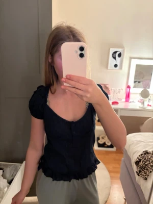 Mörkblå blus med puffärmar - Söt mörkblå blus med korta puffärmar och knappar framtill. Blusen har en figurnära passform och volangkant nedtill. Har aldrig använt. Priset kan diskuteras ❣️