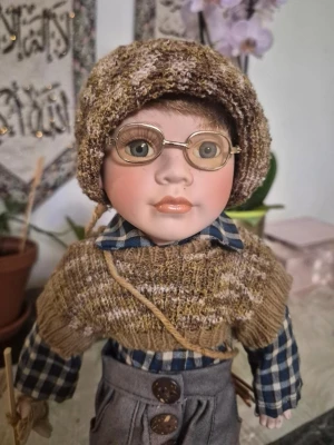 Vintage GEPPEDDO Porslin Vit Bondepojke Blå Ögon m. Glasögon Docka Storlek 45 cm - En söt docka klädd i traditionell folkdräkt med rutigt skjorta, grå byxor med knappar och lappar, samt en stickad brun sjal och matchande huvudbonad. Dockan håller en liten käpp och har svarta skor. Perfekt som prydnad eller samlarobjekt.