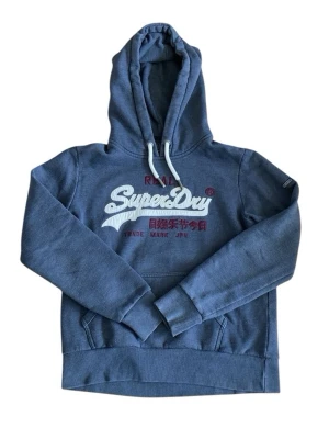 Blå Superdry hoodie - Säljer en blå hoodie från Superdry med stor broderad logga i vitt och röda detaljer på bröstet. Hoodien har huva med vita dragsnören och en klassisk känguruficka framtill. Perfekt för dig som gillar streetwear och vill ha något bekvämt och snyggt.