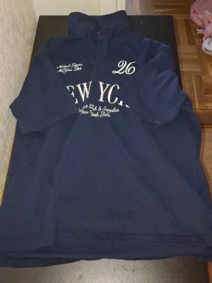 Mörkblå henley med New York-tryck - Snygg mörkblå henley med tryck på bröstet där det står 'New York' och siffran 26. Skjortan har krage, knappar framtill och långa ärmar. Perfekt för dig som gillar streetwear och vill ha en avslappnad men cool look.