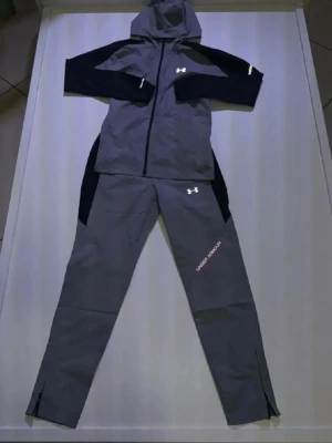 Grå träningsset från Under Armour - Snyggt grått träningsset från Under Armour med matchande jacka och byxor. Jackan har huva, dragkedja och svarta detaljer på ärmarna. Byxorna har logga och text på benet samt svarta paneler på sidorna. Perfekt för träning eller sportaktiviteter.