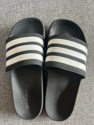 Badtofflor Adidas - Använda, men i fint skick. En spricka i sulan under, men inget som stör. Se bild. Storlek 40 1/2