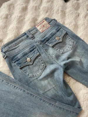 Ljusblå jeans från True Religion - Säljer ett par ljusblå jeans från True Religion joey low rise  med ikoniska sömmar och snygga fickdetaljer bak. Jeansen har normal midja, raka ben och klassisk femficksmodell. Tillverkade i mjuk denim med silverfärgade knappar och nitar. Perfekta för en avslappnad och trendig look. Storlek 24. Uppsydda och kan ha lite skavanker nertill men inte riktigt något man tänker på