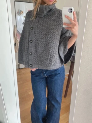 Grå stickad poncho med knappar - Säljer en grå stickad poncho med hög polokrage och stora svarta knappar framtill. Ullblandning. Funkar som en hårig jacka eller varmare kofta. 