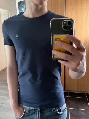 Mörkblå t-shirt från Ralph Lauren - Säljer en mörkblå t-shirt från Ralph Lauren med den klassiska loggan broderad på bröstet. T-shirten har rund hals, korta ärmar och en slim passform. Perfekt till jeans eller shorts för en clean och enkel stil.