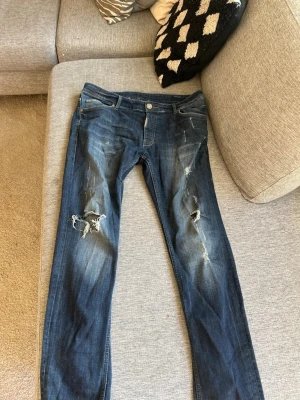 Blå slitna jeans med hål - Säljer ett par blå jeans med slitningar och hål på båda benen. Jeansen har en klassisk femficksmodell och knappgylf. Snygg tvätt med ljusa partier och råa detaljer för en trendig look.