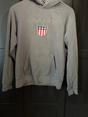 Grå hoodie från GANT med logga - Snygg grå hoodie från GANT med broderad logga och amerikanskinspirerat sköldmärke på bröstet. Klassisk känguruficka framtill och huva med dragsko. Tillverkad i mjukt bomullsmaterial, perfekt för en avslappnad stil.