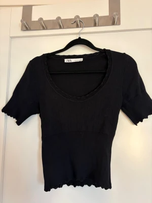 Svart ribbad topp från Zara - Snygg svart ribbad topp från Zara med korta ärmar och v-ringad hals. Toppen har vågiga kanter vid ärmslut och nederkant samt en figurnära passform. Perfekt att matcha med jeans eller kjol för en trendig look.