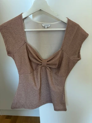 Glittrig topp med rosett  - Supersnygg glittrig topp i champagnefärg från With Black. Toppen har en stor rosettdetalj framtill, djup v-ringning och är ärmlös. Perfekt för dig som vill sticka ut lite extra med en unik och trendig look. Från bubbelroom. Helt oanvänd 
