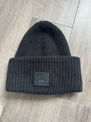 Svart mössa från Acne Studios - Säljer en svart ribbstickad mössa från Acne Studios med den ikoniska ansiktsloggan framtill. Mössan är i tjockt, mjukt material och har en bred uppvikt kant. Perfekt för kalla dagar och ger en clean, stilren look. Mössan har lite veck som jag inte kan få bort som man ser på bilderna därav billigare pris! Men den är fortfarande super fin 