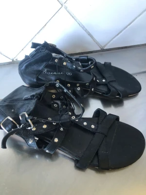 Svarta sandaler med nitar från Avenue - Snygga svarta sandaler från Avenue med flera remmar dekorerade med silverfärgade nitar och spännen. Dragkedja baktill för enkel på- och avtagning. Platt sula och öppen tå ger en cool och avslappnad look. Materialet ser ut att vara konstläder.     Har ej använt dessa helt nya med prislapp 
