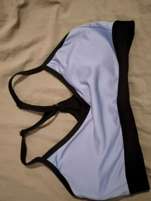 Ljuslila sport-bh med svarta detaljer - Sport-bh i ljuslila med svarta kanter och band. Bh:n har bred resår under bysten och justerbara axelband. Mjuk och stretchig modell utan bygel, perfekt för träning eller vardag. Märke saknas.