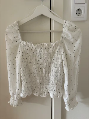 Vit prickig blus med puffärm - Supersöt vit blus med svarta prickar och puffiga långärmar. Smockad över bröstet och midjan för en tight och bekväm passform. Rynkade detaljer vid ärmslut och fyrkantig ringning ger en romantisk vibe. Storlek 34 i nyskick!