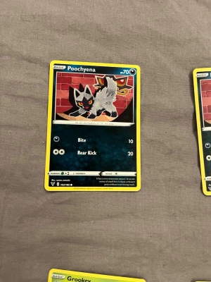Pokémonkort Poochyena - Säljer ett Pokémonkort med Poochyena-motiv. Kortet har en svart och grå tecknad figur mot en röd bakgrund med gula detaljer. Kortet är rektangulärt och gjort av papper/kartong. Perfekt för samlare eller Pokémon-fans.