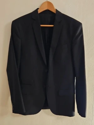 Svart kavaj från Filippa K - Stilren svart kavaj med smal passform och två knappar framtill. Klassisk design med spetsiga slag och två fickor. Perfekt för en dressad look. Materialet känns slätt och lätt, troligen polyester eller ullmix.