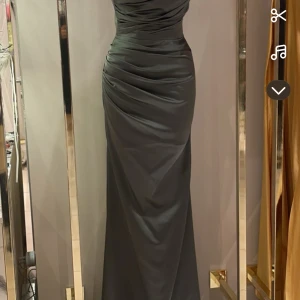 Mörkgrön långklänning - Elegant mörkgrön långklänning med draperad design och smickrande passform. Klänningen har en vacker omlott-effekt och är perfekt för formella tillfällen. Tillverkad i ett glansigt material som ger en lyxig känsla. Midjan är insydd samt släp baktill .  Inköpspris 5800kr. Kommer med egna tygpåsa!! Den är använd några timmar på en fest endast 