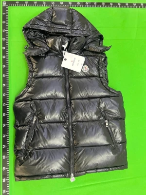 Svart dunväst  - Snygg svart dunväst från Moncler med glansig finish och avtagbar huva. Västen har två dragkedjeförsedda fickor fram och klassisk Moncler-logga på bröstet. Perfekt för lager-på-lager och håller dig varm under kyliga dagar.
