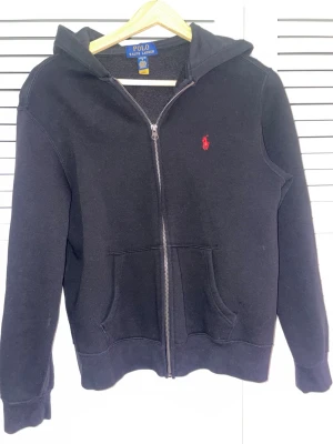 Svart hoodie från Polo Ralph Lauren - Svart hoodie från Polo Ralph Lauren med dragkedja framtill och klassisk röd logga på bröstet. Tröjan har huva, två fickor fram och ribbade muddar. Perfekt för en chill och stilren look. Materialet känns mjukt och bekvämt.
