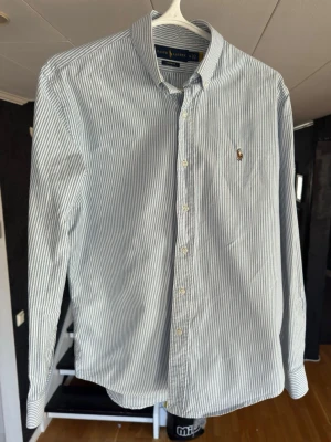 Randig skjorta från Ralph Lauren - Klassisk blå- och vitrandig skjorta från Ralph Lauren med button-down krage och broderad logga på bröstet. Skjortan har långa ärmar och knäppning framtill. Perfekt för en stilren och tidlös look.