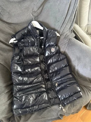 Svart glansig dunväst från Moncler - Snygg svart dunväst från Moncler med glansig finish och hög krage. Västen har två dragkedjeförsedda fickor framtill och en Moncler-logga på bröstet. Perfekt för lager på lager och streetstyle.