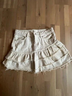 Beige volangkjol med råa kanter - Supersöt vit kortkjol med volanger från Gina. Kjolen har fickor fram och bälteshällor i midjan. Jättefin till sommaren! Kjolen har en normal passform.💕