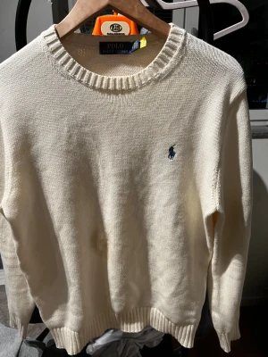 Vit stickad tröja från Polo Ralph Lauren - Klassisk vit stickad tröja från Polo Ralph Lauren med rund halsringning och diskret blå logga broderad på bröstet. Tröjan har ribbade muddar vid ärmslut och nederkant. Perfekt för en clean och stilren look.