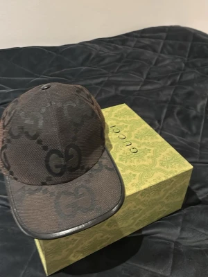 Svart keps med GG-mönster från Gucci - Säljer en svart keps från Gucci med det klassiska GG-monogrammet i mörkgrått över hela kepsen. Kepsen har böjd skärm och justerbar rem bak. Materialet är slitstark canvas och detaljerna är riktigt snygga. Perfekt för dig som gillar exklusiva accessoarer. Kvitto och box finns, beställd på guccis hemsids