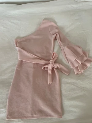 Ljusrosa oneshoulder klänning  - Säljer en ljusrosa kortklänning från Boohoo med oneshoulder-design och volangärm. Klänningen har ett brett knytband i midjan som ger en snygg siluett. Materialet är mjukt och strukturerat🌸