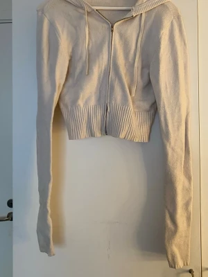 Vit cropped hoodie med dragkedja - Säljer en vit cropped hoodie med dragkedja framtill och ribbad nederkant. Hoodien har långa ärmar, huva med snören och är stickad i mjukt material. Perfekt för dig som vill ha en chill och trendig look.