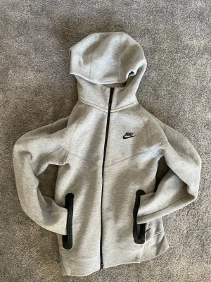 Grå Nike hoodie med dragkedja - Säljer en grå hoodie från Nike med hel dragkedja och svart logga på bröstet. Hoodien har två svarta fickor med dragkedja och en stor huva. Materialet är mjukt och stretchigt, perfekt för chill eller träning.