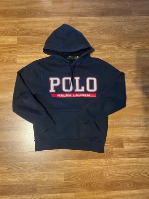 Mörkblå hoodie från Polo Ralph Lauren - Snygg mörkblå hoodie från Polo Ralph Lauren med stor logotyp i vitt och rött på bröstet. Klassisk huva med snörning och ribbade muddar. Perfekt för dig som vill ha en lite mer ”grisch” look . Materialet är mjuk bomull. 